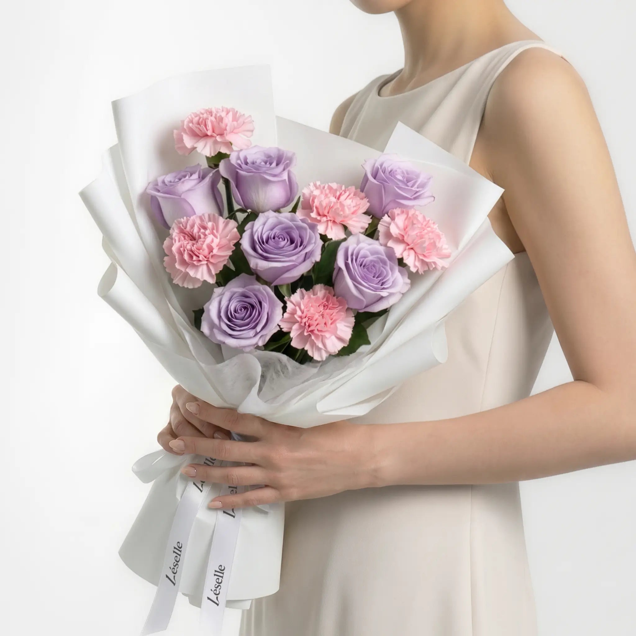Bouquet of lavender roses and blush carnations held by a person in a white dress on a white background